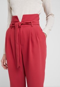 Vysoké červené kalhoty s širokými nohavicemi, pásek z látky na uvázání, sklady v pase a boční kapsy. Měkký, texturovaný materiál.