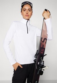 Bijela dugoslojna flis majica s polu-zipom i zig-zag uzorkom. Crni detalji i uski kroj. Ski oprema u desnoj ruci.