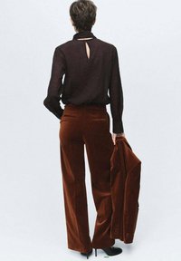 Langsleeve donkere blouse met een uitsnede op de rug, gecombineerd met hoog-geplaatste roestkleurige fluwelen broek. Model houdt een bijpassende fluwelen jacket vast. Zwarte hakken.