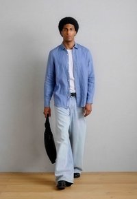 Άνδρας με αφρομάλλες, που φοράει μπλε πουκάμισο, λευκή μπλούζα, φαρδιά ανοιχτόχρωμα τζιν, μαύρα παπούτσια και κρατάει μια μαύρη τσάντα, περπατώντας σε ξύλινο πάτωμα.