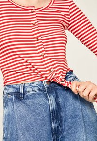 Maglietta a righe rosse e bianche a maniche lunghe con dettagli di bottoni, legata in vita, abbinata a jeans in denim blu sbiadito con chiusura frontale a bottoni.