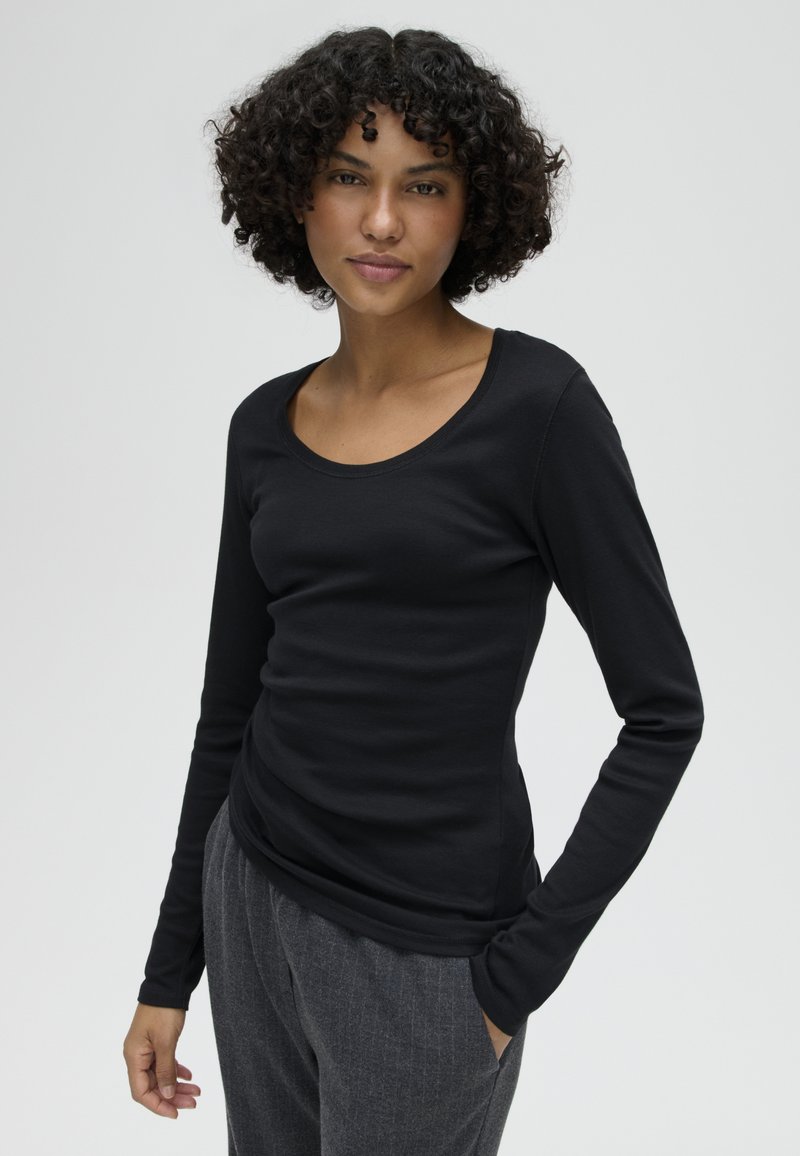 Vrouw met krullend haar, gekleed in een zwarte top met lange mouwen en een grijze broek, staat met één hand in haar zak tegen een effen achtergrond.