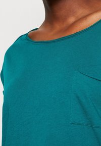 T-shirt a maniche corte teal con scollatura rotonda e dettaglio tasca sul lato sinistro. Tessuto di cotone liscio con una leggera texture.