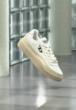 Weißer Diesel-Sneaker mit beiger Sohle, der über einem polierten Boden, einer Glassteinwand und Betonpfeilern im Hintergrund schwebt.