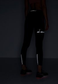 Leggings neri con strisce laterali riflettenti e logo, realizzati in materiale elastico. Indossati con sneakers nere caratterizzate da un suola prominente.