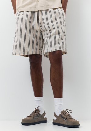 Mann trägt beige Shorts mit schwarzen vertikalen Streifen, weiße Kniestrümpfe und braune Schnürschuhe mit offenem Absatz.