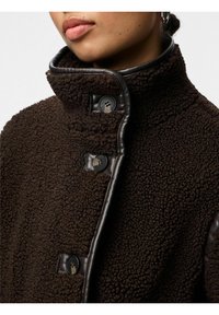 Rjava flis jakna z visokim ovratnikom, ki ima temne usnjene obrobe, štiri črne gumbe in teksturirano površino.