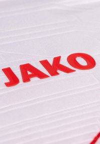 Maglia sportiva bianca con tessuto testurizzato, con il logo rosso "JAKO" in evidenza. Design semplice e moderno.