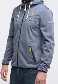 Grå zip-up hoodie med dragsko på huvan, två zip-fickor med gula detaljer och broderad logotyp på bröstet. Slät textur.