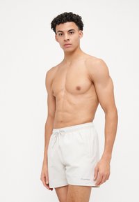Baltos maudymosi šortai su elastine juosta ir virvele. Kairėje apatinėje pusėje yra mažas Calvin Klein logotipas. Lygus paviršius, minimalus dizainas.