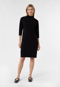 Schwarzes knielanges Kleid mit hohem Kragen und dreiviertel Ärmeln, aus glattem Stoff, getragen mit schwarzen Schuhen. Einfaches, elegantes Design.