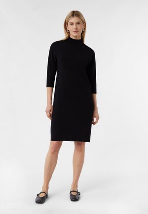 Robe noire midi avec un col montant et des manches trois-quarts, fabriquée en tissu lisse, portée avec des chaussures noires. Design simple et épuré.