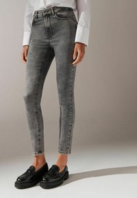 Dopasowane czarne dżinsy z wyblakłym szarym odcieniem, pięć kieszeni, średni stan, w tonacji przeszycia, oraz zwężana nogawka, w połączeniu z błyszczącymi czarnymi mokasynami.