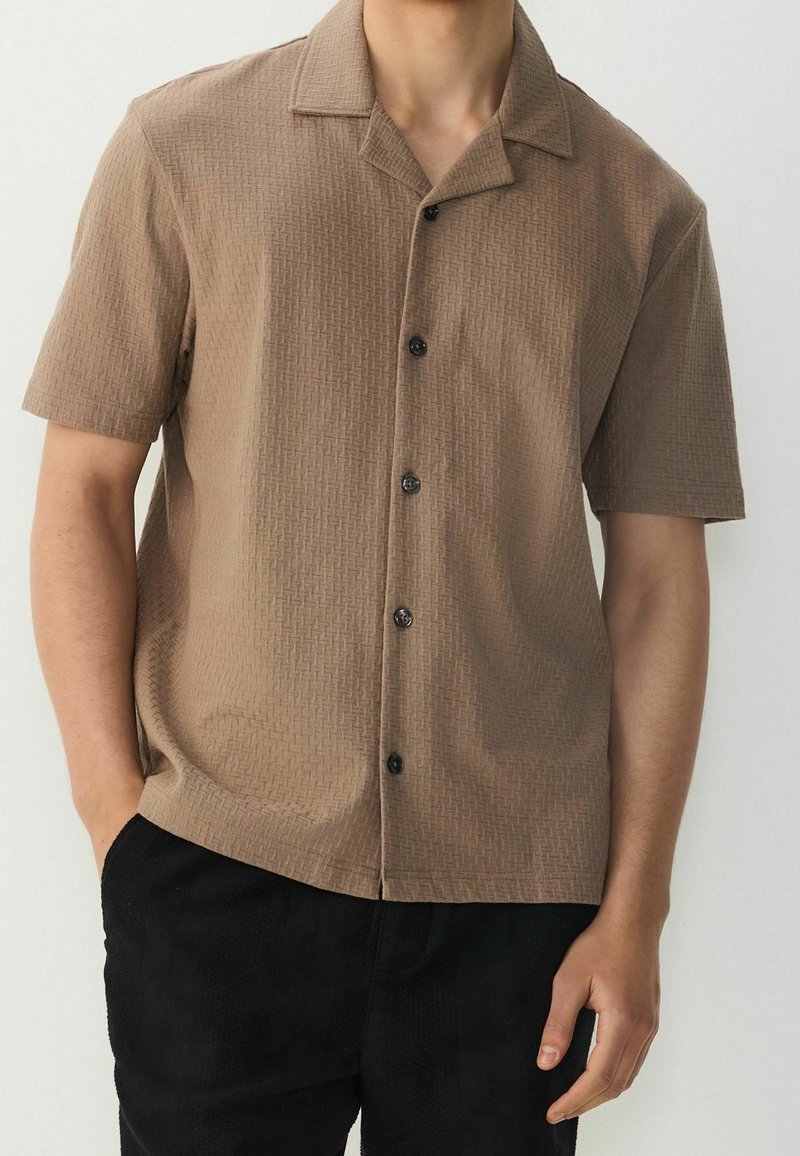 Homme portant une chemise beige à manches courtes texturée avec un col cranté et un pantalon noir, debout devant un fond neutre.