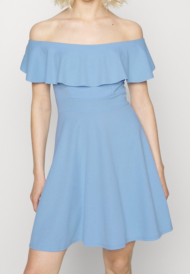 Robe bleu clair décolletée sur les épaules avec un col volanté. Corsage ajusté menant à une jupe évasée en tissu lisse et extensible.