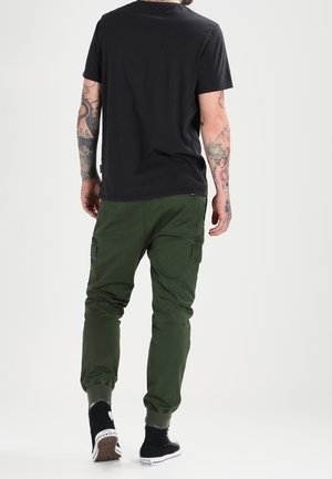 Homme aux bras tatoués portant un t-shirt noir, un pantalon cargo vert et des baskets montantes noires, debout dos tourné vers l'avant.