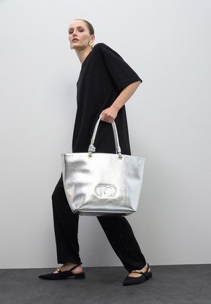 Srebrna metalik torba za nošenje s logotipom, s dva ručka i glatkom teksturom, u kombinaciji s opuštenom crnom odjećom i crnim cipelama.
