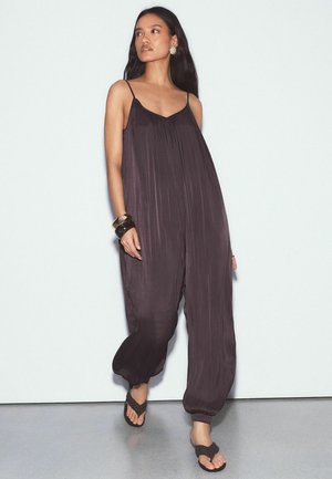 Moteris dėvi laisvą tamsiai violetinės spalvos rožinių dirželių kombinezoną be rankovių, juodus sandalais, dideliais auskarais ir masyviais apyrankėmis.