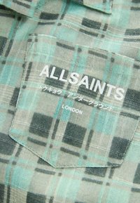 Grön och grå-rutig skjorttyg med en texturerad väv; har en ficka med "ALLSAINTS"-logotyp och japansk text.