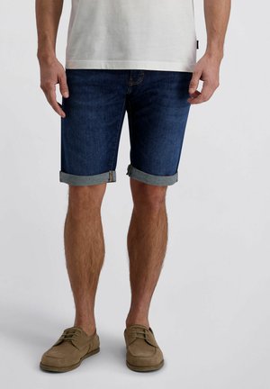 Homme portant un short en jean bleu foncé retroussé, un t-shirt blanc et des chaussures marron à lacets, debout devant un fond uni.