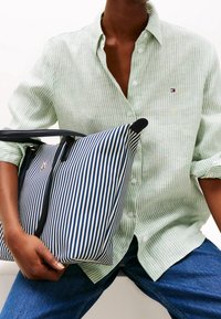 Chemise rayée verte et blanche avec des boutons, associée à un grand sac fourre-tout rayé bleu marine et blanc avec des poignées noires et un détail logo doré.