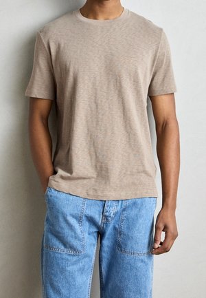 Man draagt een beige T-shirt met korte mouwen en ronde hals en lichtblauwe spijkerbroek, staand tegen een effen lichtgrijze achtergrond.