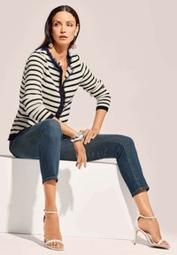 Streepjessende gebreide cardigan in crème en marineblauw, met franjes, gecombineerd met donkerblauwe jeans en witte hakken met bandjes. Zilveren accessoire om de pols.