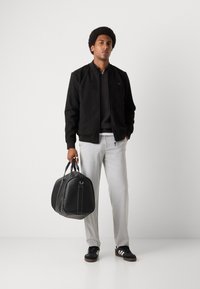 Black suede bomber jacket over a dark shirt, light grey pants, and black sneakers. Holding a black leather duffel bag with white stitching.