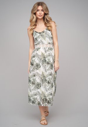 Ung kvinde med langt blond hår iført en hvid midi kjole med grønne tropiske blade og blomsterprint og sandaler, stående mod grå baggrund.