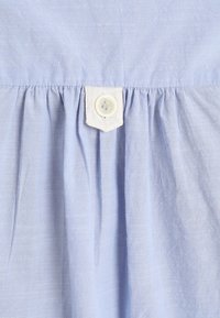 Tissu bleu clair avec tissage texturé et plis froncés maintenus par un onglet blanc cousu avec un bouton rond blanc central.