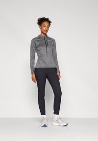 Grå langermet zip-up topp med høy krage, parret med svarte tettsittende bukser og lys grå joggesko med oransje detaljer.