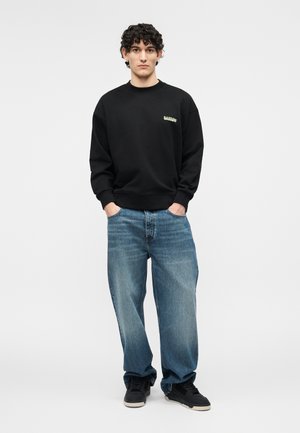 Jeune homme debout, mains dans les poches, portant un sweat-shirt noir avec un logo jaune, un jean bleu ample et des baskets noires sur un fond neutre.