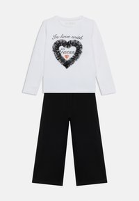 Λευκό πουκάμισο με μακριά μανίκια με το κείμενο "Ερωτευμένη με Guess", μαύρη καρδιά με βολάν, κόκκινη καρδιά με γκλίτερ, συνδυασμένο με μαύρο παντελόνι φαρδιάς γραμμής.