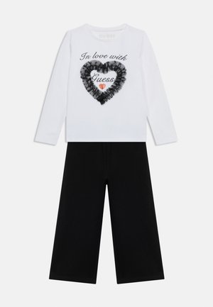 Balta krekla ar gariem piedurknēm ar tekstu "In love with Guess", melnu kroku sirdi un sarkanu mirdzošu sirdi, kopā ar melnām plašām biksēm.