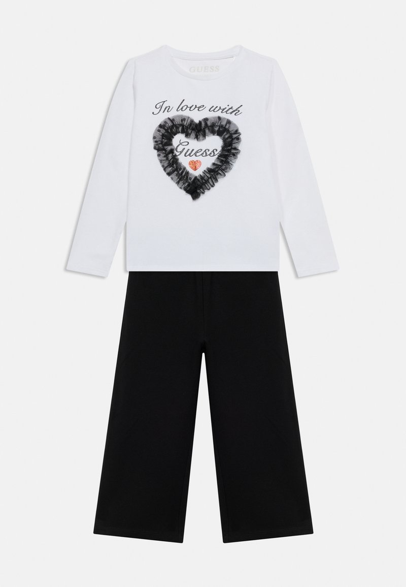 Λευκό πουκάμισο με μακριά μανίκια με το κείμενο "Ερωτευμένη με Guess", μαύρη καρδιά με βολάν, κόκκινη καρδιά με γκλίτερ, συνδυασμένο με μαύρο παντελόνι φαρδιάς γραμμής.