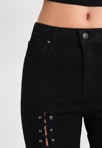 Černé denimové džíny s vysokým pasem, předními kapsami a dekorativními šněrovacími prvky s očičkami na bočních švech. Hladká textura.