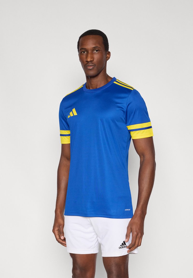 Maglietta atletica blu con strisce gialle sulle maniche, realizzata in materiale liscio. Presenta un logo sul petto a sinistra e dettagli a contrasto. I pantaloncini sono bianchi.