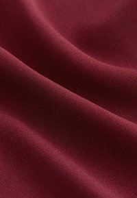 Burgundy fabric with a smooth, matte texture, displaying subtle ripples and folds. The surface appears finely woven and uniform in color.