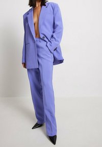 Lila Anzugset mit einem taillierten Blazer mit einem einzigen Knopf und weit geschnittenen Hosen. Der Stoff hat eine glatte Textur; schwarze spitze Schuhe.
