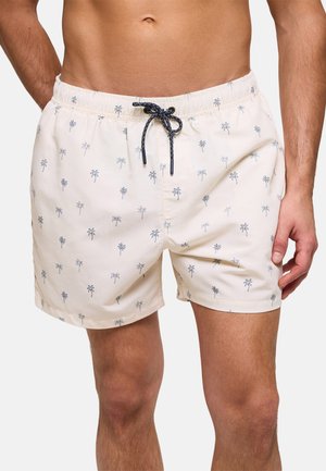 Hombre con pantalones de baño color crema con pequeños estampados oscuros de palmeras y un cordón negro atado en la cintura.