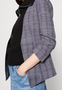 Veste à carreaux grise avec des accents blancs ; tissu léger, devant ouvert et manches retroussées. Portée sur un pull noir et un jean bleu.