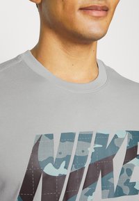Gray t-shirt featuring a large, multi-colored blue camouflage Nike logo, round neckline, and a smooth fabric texture.