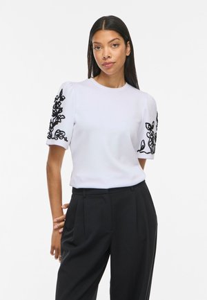 Femme aux longs cheveux foncés portant un haut blanc à manches courtes avec une broderie florale noire sur les manches et un pantalon noir taille haute.