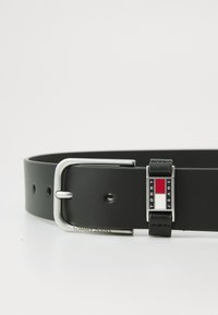 Zwarte leren riem met een zilveren gesp, voorzien van een rechthoekig Tommy Jeans-logo in rood, wit en blauw. Gladde textuur, minimalistisch ontwerp.