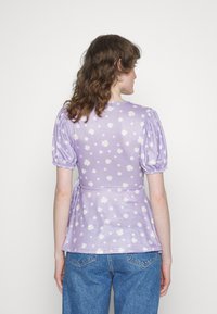 Blusa lavanda com mangas bufantes, apresentando um padrão de margaridas, cintura peplum e textura suave. Combinada com jeans de ganga azul.