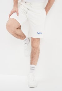 Witte katoenen shorts met een tailleband met trekkoord, voorzien van zijzakken en een blauwe geborduurde "Creek" logo. Gekomplementeerd met witte sneakers en gestreepte sokken.