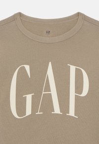 Béžové bavlněné tričko s kulatým výstřihem. Na přední straně se nachází velké bílé logo "GAP". Hladký materiál, volný design pro děti.