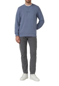 Muž oblečený v modrom mikině, šedých džínsoch a bielych teniskách, stojaci s jednou rukou za chrbtom na bielom pozadí.