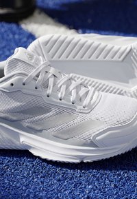 Paire de chaussures de sport blanches avec semelles texturées reposant sur une surface bleue, montrant un tissu en mesh et des détails de lacets.