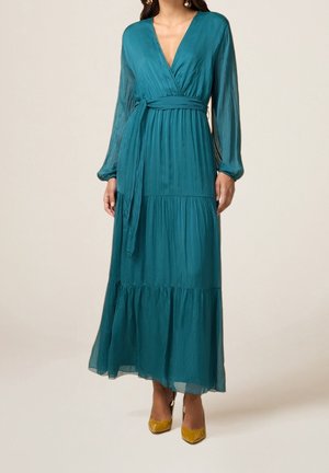 Robe portefeuille à manches longues couleur sarcelle avec jupe à volants et ceinture à la taille, associée à des escarpins jaunes moutarde brillants à bouts pointus sur fond neutre.